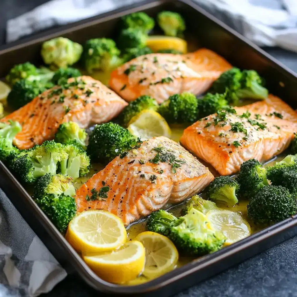 A baking tray with four salmon fillets surrounded by lemon slices and broccoli florets.