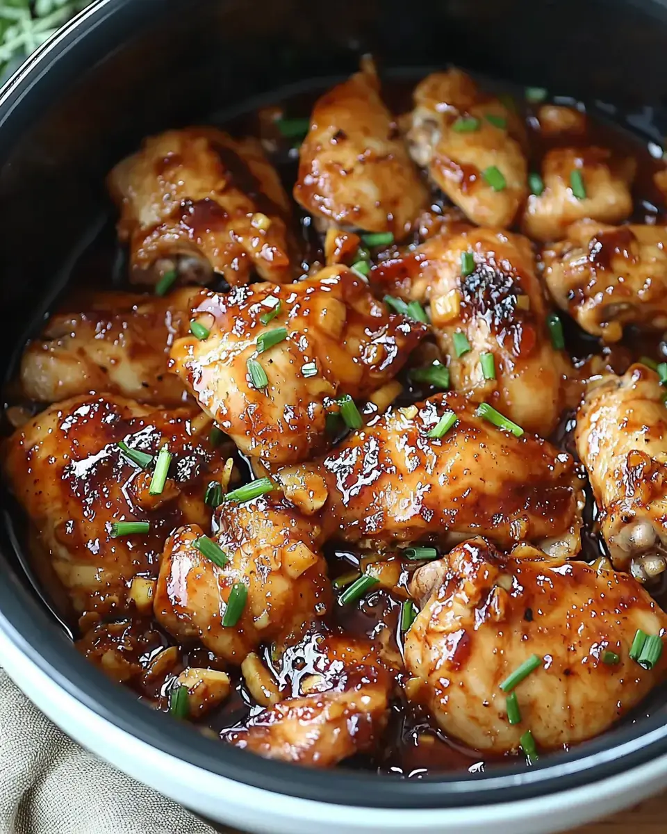 A close-up view of cooked chicken thighs glazed in a shiny brown sauce and garnished with chopped chives in a black bowl.