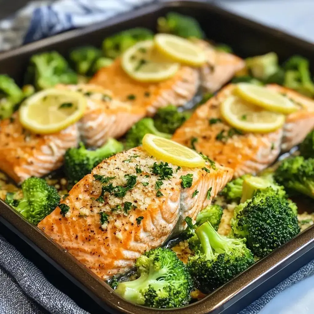 Three baked salmon fillets topped with lemon slices and surrounded by broccoli florets in a roasting pan.