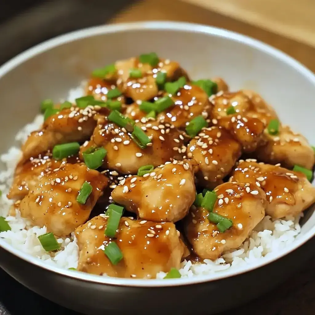 A bowl of chicken pieces glazed with sauce, garnished with green onions and sesame seeds, served over a bed of white rice.