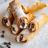 A plate of filled cannoli dusted with powdered sugar, featuring a creamy filling with chocolate pieces inside.