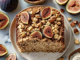 A slice of moist fig cake topped with fresh figs and chopped nuts, surrounded by whole figs and nuts on a marble surface.