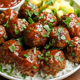 A plate of meatballs covered in sauce and garnished with chopped herbs, served over rice with a side of diced fruit and sauce.