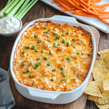 A creamy, cheesy baked dish topped with green onions is served alongside celery sticks, carrot sticks, and tortilla chips.